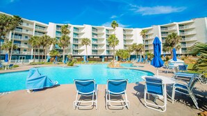 A hotel pool area with multiple lounge chairs and umbrellas, surrounded by palm trees and a large building with balconies. - Sandcastle Condominium Unit 128 (Port Aransas)