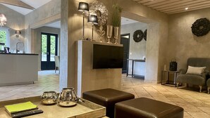 Een moderne woonkamer met een houten salontafel, leren poefs, een aan de muur bevestigde televisie, en een keuken die door een open deur zichtbaar is.