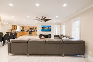 A modern living room with a sectional sofa, a dining area with a table and chairs, a kitchen with wooden cabinets, and a ceiling fan.