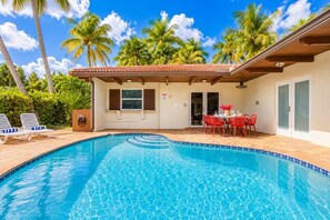 A swimming pool with a tiled edge, surrounded by lounge chairs, a dining area with red chairs, and palm trees in the background.