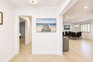 Un couloir orné d’un tableau représentant une plage et une jetée, un coin repas avec des chaises et une table, et une vue sur l’océan par une fenêtre.