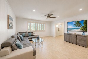 Een ruime woonkamer met een hoekbank, een glazen salontafel, een plafondventilator, een grote flatscreen-tv en uitzicht op het strand.