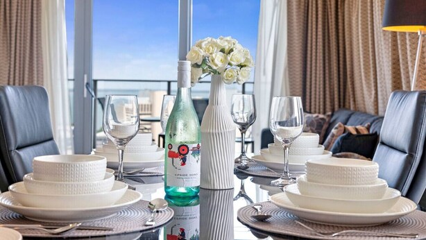 A dining table set for a meal with white plates, a bottle of wine, and a vase of white flowers.