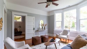 A living room with a ceiling fan, a sofa, a chair, a coffee table, and a window with a view of the outside. - Bright & Cozy Home | Walkable, Private Yard + WiFi (Seattle)