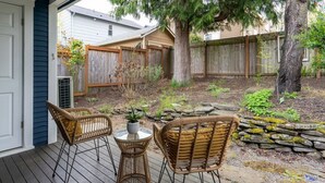 A wooden deck with wicker chairs and a small table, surrounded by a garden with a stone wall and a wooden fence. - Bright & Cozy Home | Walkable, Private Yard + WiFi (Seattle)