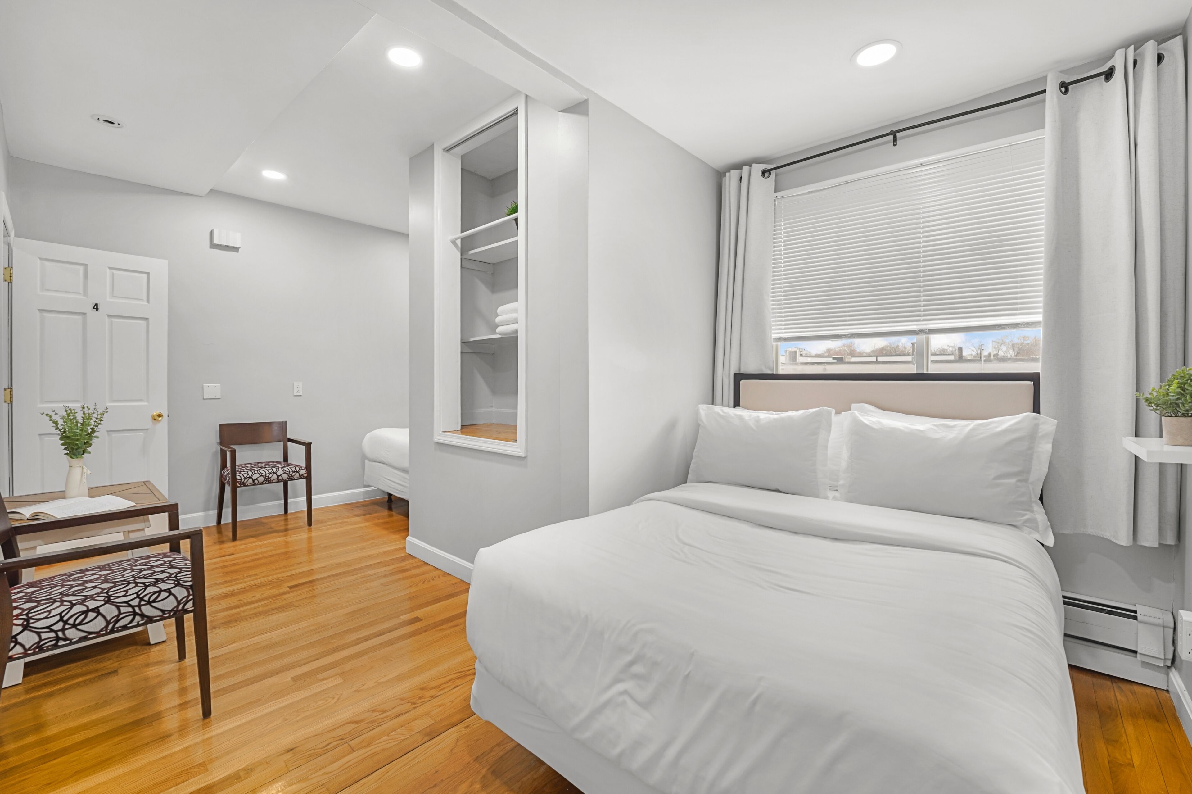 A bedroom with a large bed, a wardrobe, a desk with a chair, and a window with blinds.