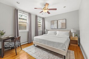A bedroom with a bed, a desk, a chair, a ceiling fan, and two windows with curtains.