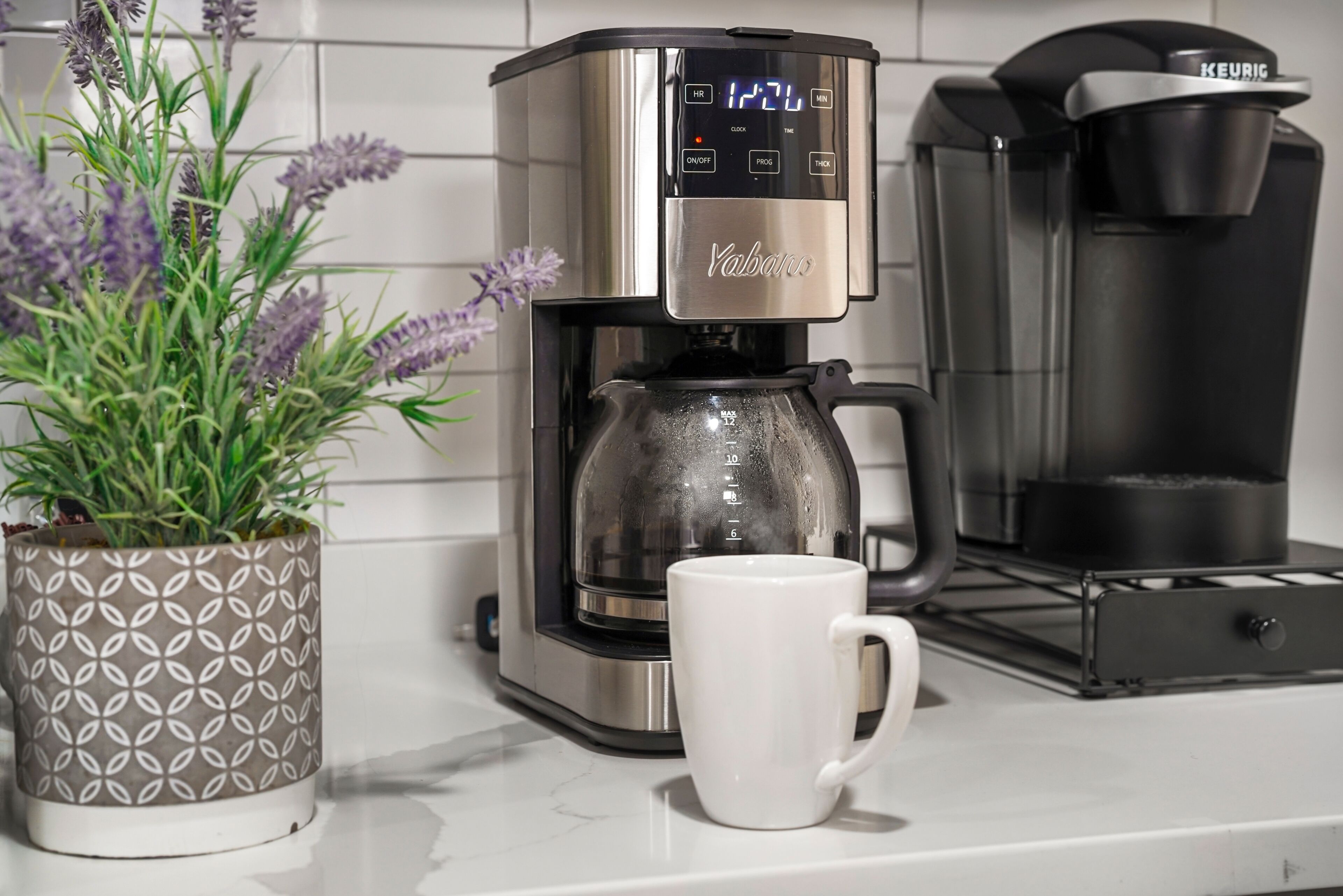 A coffee maker with a digital display, a white mug, and a potted plant on a kitchen counter.