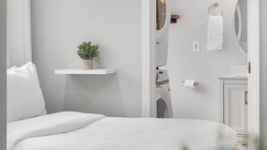 Een slaapkamer met een bed, een kleine potplant, een witte muur en een deur naar een badkamer.