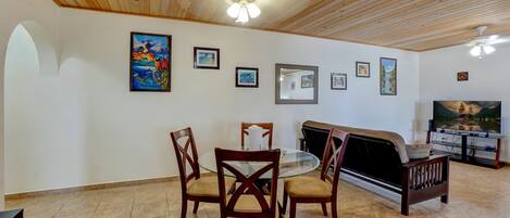A dining area with a round glass table, four wooden chairs, a wooden bench, and a television on a stand.