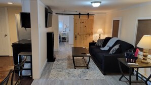 A living room with a black sofa, a coffee table, and a TV mounted on the wall. There is a wooden door in the background leading to another room. - Beautiful lake side cottage (Hampden)