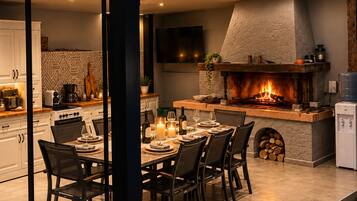 An outdoor dining area with a stone fireplace, a kitchen in the background, and a modern design.