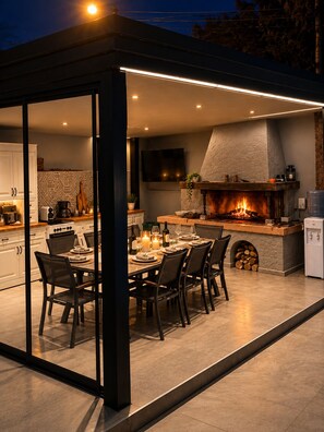 An outdoor dining area with a stone fireplace, a kitchen in the background, and a modern design. - Joan Luxury Villa Apt with private pool & Gym (Tirana)
