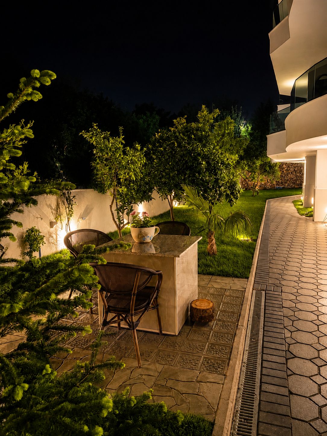 An outdoor patio area with a stone table and chairs, surrounded by lush greenery and a well-lit pathway.