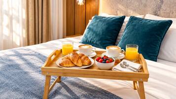 Un plateau de petit-déjeuner bien présenté, avec des croissants, un bol de fruits et deux tasses de café, posé sur un lit doté d’oreillers bleus et d’une tête de lit en bois.