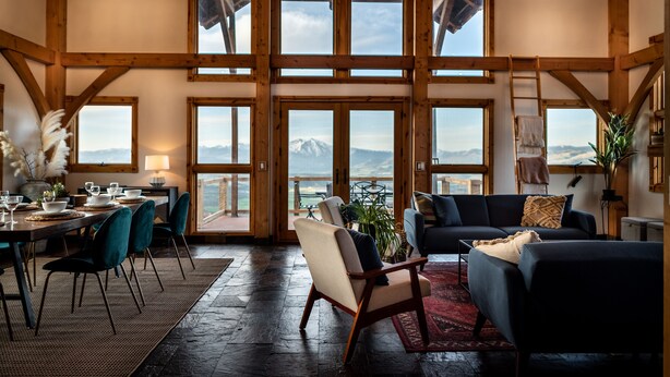 Ein geräumiger Raum mit einer großen Holzbalkendecke, zahlreichen Fenstern mit Blick auf die Berge, einem Essbereich mit Tisch für vier Personen und einer gemütlichen Sitzecke mit Sofa und Sesseln.