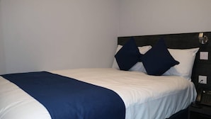 A neatly made bed with a white and blue bedspread, two dark blue pillows, and a bedside table with a phone. - Blue Sapphire Hotel (Ilford)