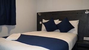 A neatly made bed with white and navy bedding, a dark headboard, and a small bedside table with a lamp. - Blue Sapphire Hotel (Ilford)