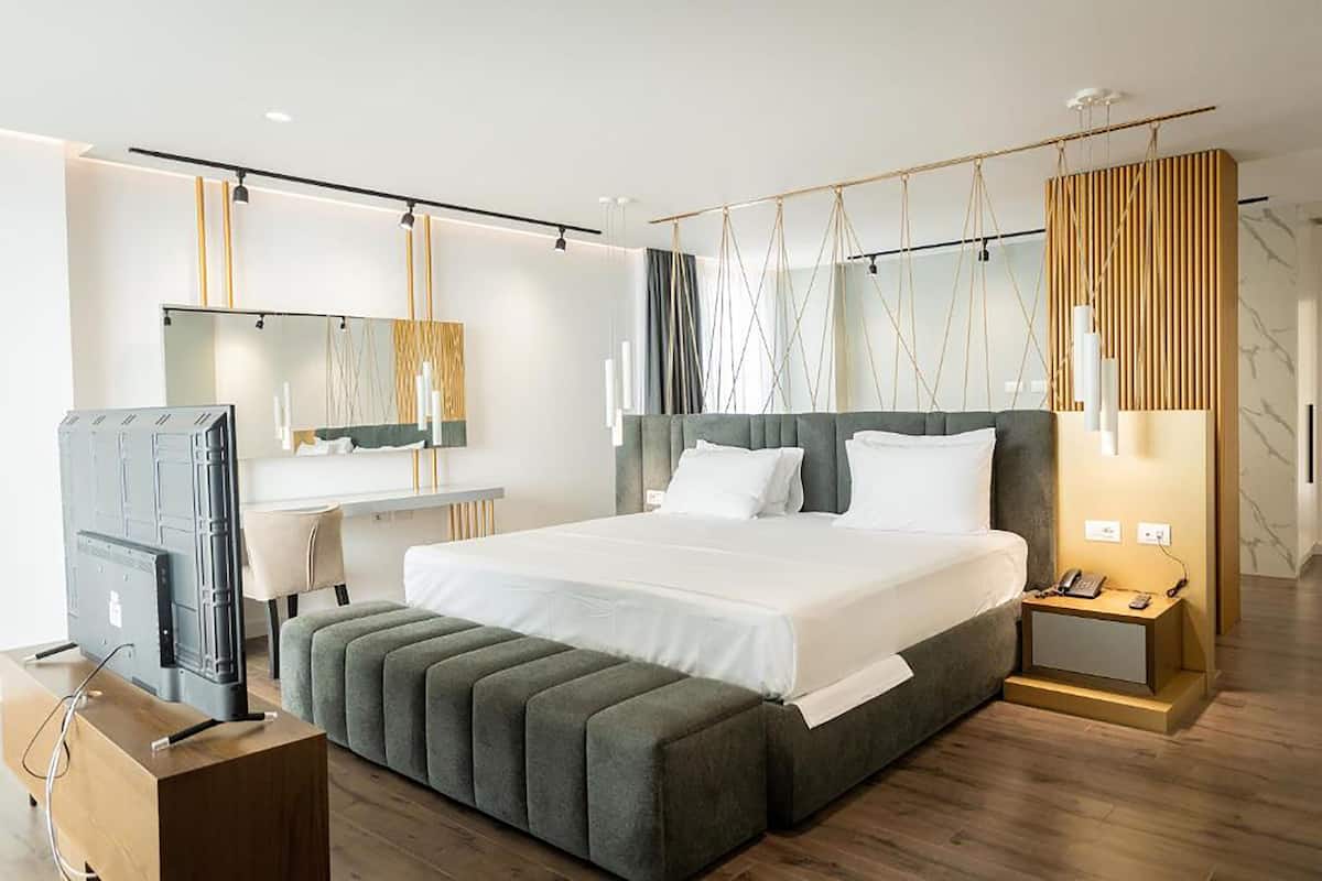 A modern hotel room with a large bed, a bedside table with a phone, a TV, and a wooden desk.