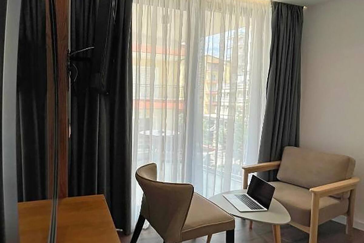 A hotel room with a bed, a desk with a laptop, a chair, a TV, and a window with curtains.