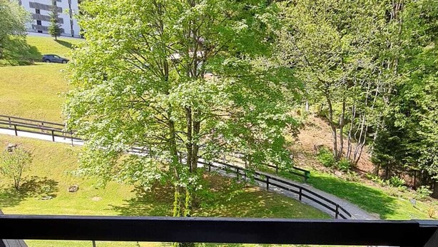 Un balcon avec deux chaises blanches et une petite table ronde, donnant sur un espace verdoyant avec des arbres et une route.