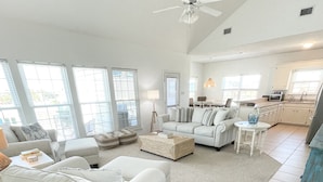 A spacious living room with a high ceiling, large windows, and a kitchenette in the background.