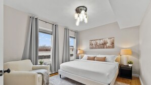 A bedroom with a bed, a chair, a nightstand, a lamp, a painting, and a view of the outside.