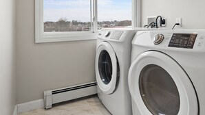 A laundry room with two front-loading washing machines, a window with blinds, a wall-mounted electrical outlet, and a tiled floor.