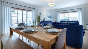 Ein Essbereich mit einem Holztisch, gedeckt für vier Personen, einem blauen Sofa und Blick ins Freie durch große Fenster.