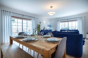Een eetgedeelte met een houten tafel voor vier personen, een blauwe bank en een uitzicht op het omliggende landschap door grote ramen.