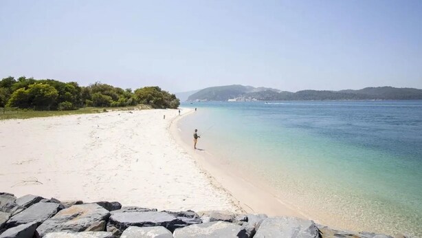 Una playa con agua cristalina, una orilla de arena y afloramientos rocosos.