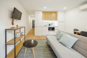 Un espace de vie moderne et compact comprenant un canapé, une petite table, un téléviseur fixé au mur et un coin cuisine.