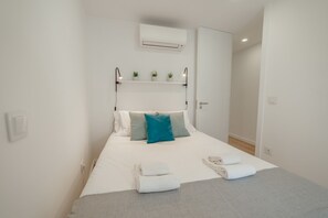 Cama bem arrumada com almofadas brancas e azul-turquesa, manta cinza e toalhas brancas dobradas sobre um cobreleito branco.