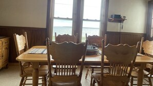 Een eetruimte met een houten tafel en stoelen, een kroonluchter en uitzicht op het water door grote ramen.
