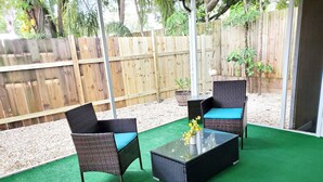 Un patio al aire libre con dos sillas de mimbre y una mesa de centro de cristal, rodeado de un cerco de madera y vegetación.
