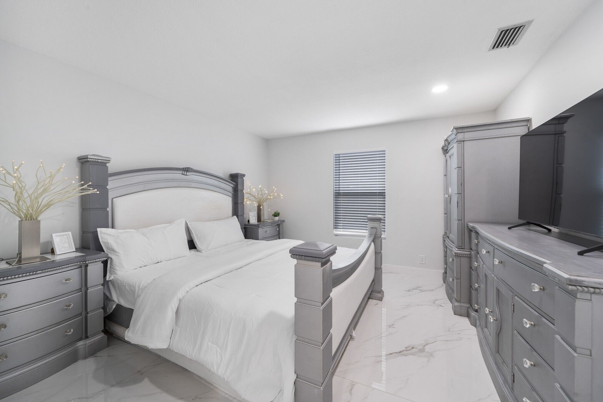 A bedroom with a large bed, a dresser, a television, and a window with blinds.