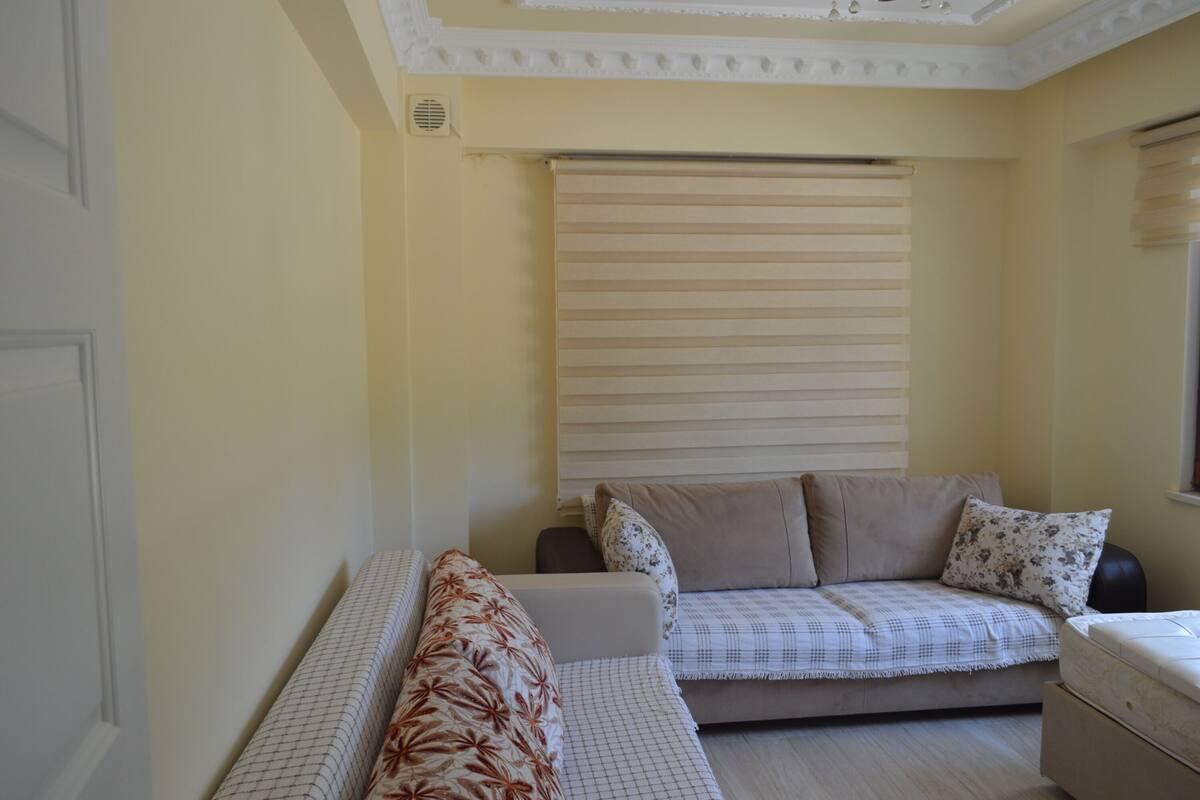 Um quarto pequeno e acolhedor, com sofá e cama, paredes bege e uma janela com persianas.