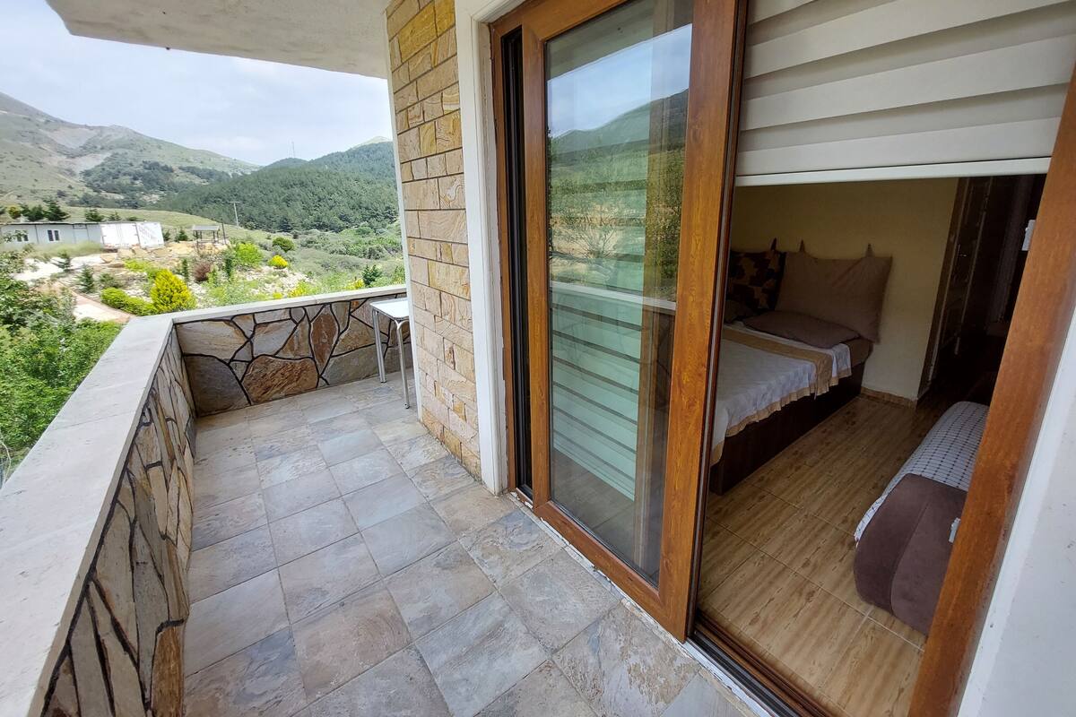 Varanda com parede de pedra e porta de madeira que dá para um quarto com cama e uma pequena mesa.