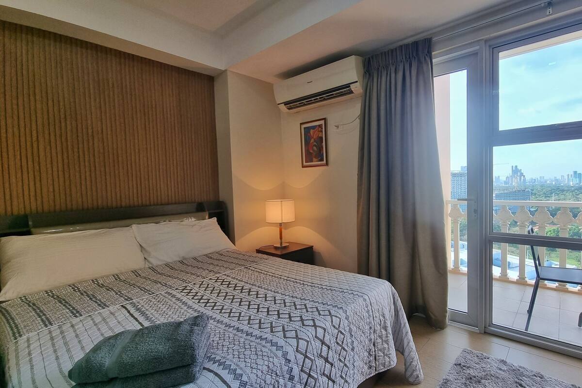 Hotellihuone, jossa on suuri sänky, yöpöytä lampun kanssa, ilmastointilaite, ikkuna, josta on näkymä kaupunkiin, ja parveke, jossa on tuoleja.