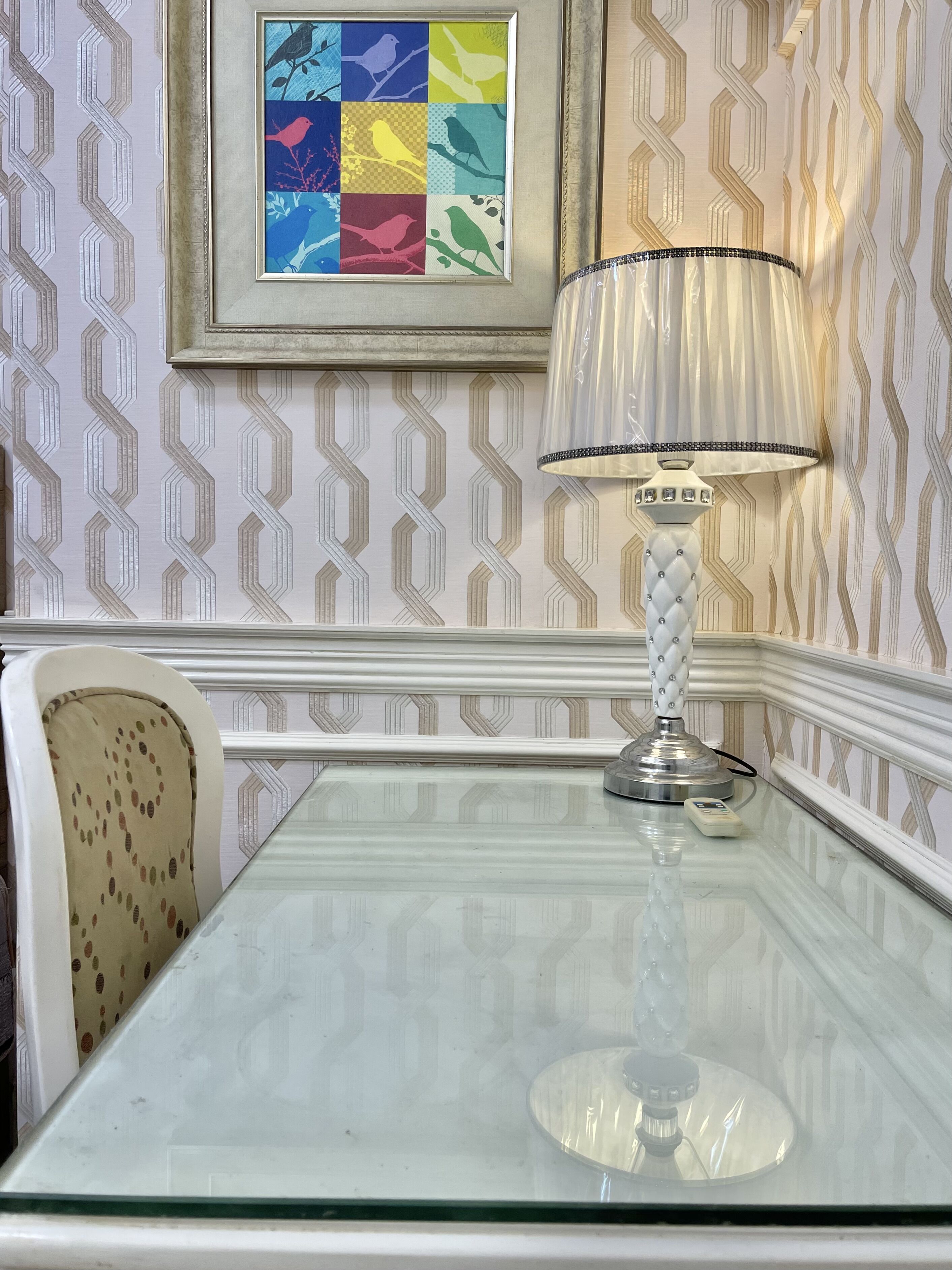 Una mesa con superficie de vidrio, una lámpara blanca y una obra de arte enmarcada encima.