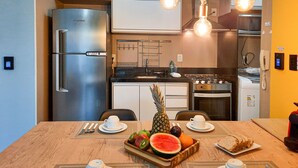 A modern kitchen with a stainless steel refrigerator, a gas stove, and a microwave. There is a dining table with fruit and bread. - Beach Way Moderno Pé na Areia 2 quartos 5 pessoas By DM Apartments (Aquiraz)