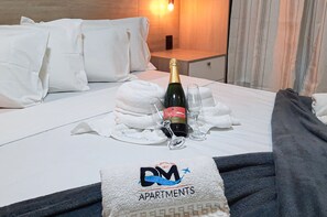 A hotel room with a neatly made bed, a bottle of champagne, and glasses on the bed. There is a towel with the hotel's logo on the bed. - Beach Way Moderno Pé na Areia 2 quartos 5 pessoas By DM Apartments (Aquiraz)