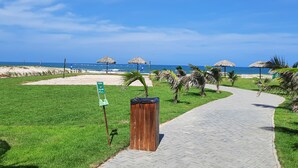 A paved pathway leading to a beach area with thatched umbrellas and palm trees. - Beach Way Moderno Pé na Areia 2 quartos 5 pessoas By DM Apartments (Aquiraz)