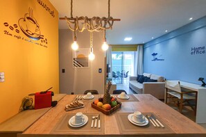 A dining area with a wooden table set for four, fruit and bread, and a coffee machine. - Beach Way Moderno Pé na Areia 2 quartos 5 pessoas By DM Apartments (Aquiraz)