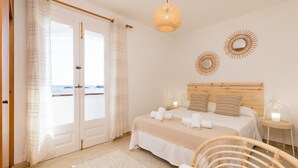A bedroom with a large bed, a wooden headboard, two wall sconces, and a view of the sea through a door. - Binilaya - Charming 3-bedroom house - seafront (Binibèquer Nou)
