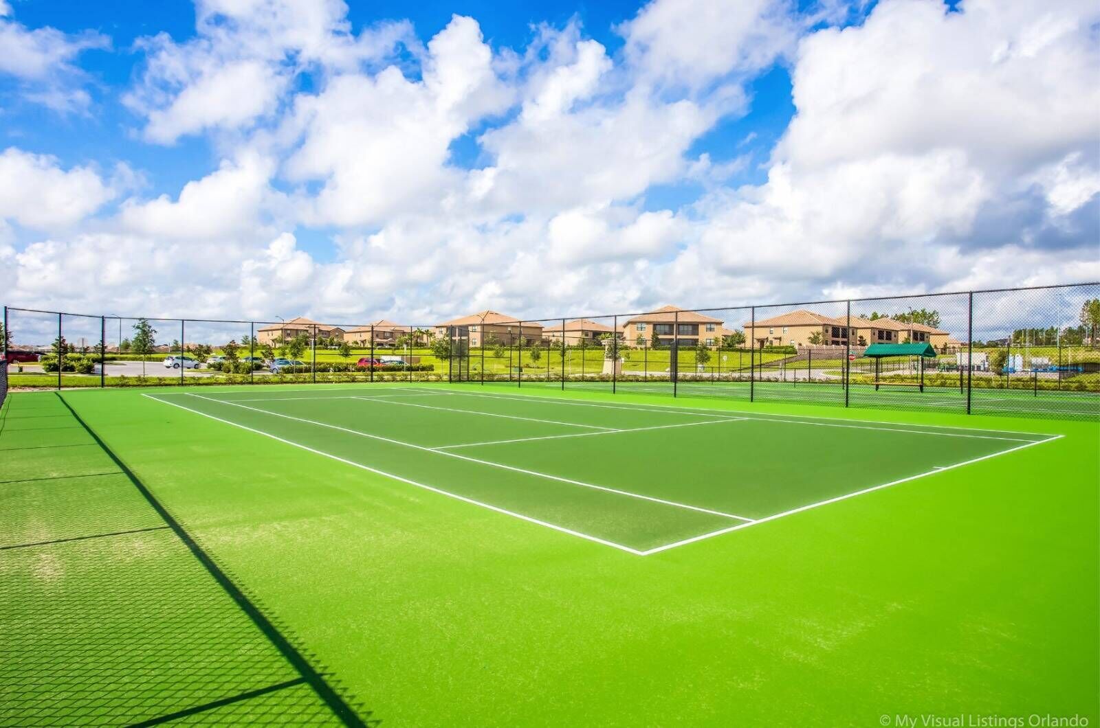 Un court de tennis avec une surface verte, entouré d’une clôture, et des immeubles d’habitation en arrière-plan.