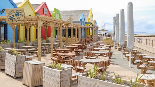 A beachside café with wooden tables and chairs, colorful buildings in the background, and a clear view of the beach. - Beeches Bach (Prestatyn)