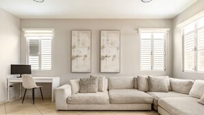 Un salon comprenant un canapé d’angle beige, un bureau blanc avec une chaise et deux œuvres d’art abstraites accrochées au mur.