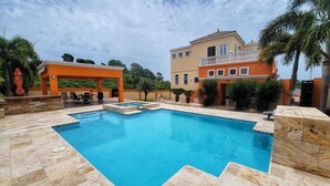 Une piscine entourée d’une terrasse en pierre, elle-même bordée d’un espace de détente couvert et d’un bâtiment à deux étages doté d’un balcon.