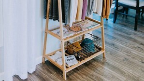 Ein Holzkleiderständer mit verschiedenen Kleidungsstücken, ein kleines Holzregal mit gefalteter Kleidung darunter und ein dekoratives Wandbild darüber.